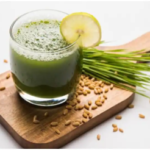 Wheatgrass juice in glass with a lemon wedge sitting on cutting board with wheatgrass seeds and wheatgrass