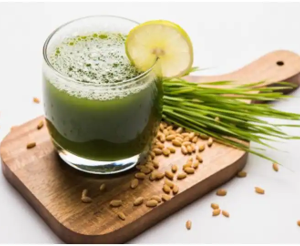 Wheatgrass juice in glass with a lemon wedge sitting on cutting board with wheatgrass seeds and wheatgrass